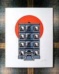 An 8"x10 art print featuring an illustrated NYC tenement building with a fire escape, black cat on the rooftop, BROOKLYN written above the doorway and a large orange-red sun behind it, photographed flat on a plaid fabric background.