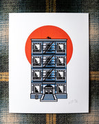An 8"x10 art print featuring an illustrated NYC tenement building with a fire escape, black cat on the rooftop, BROOKLYN written above the doorway and a large orange-red sun behind it, photographed flat on a plaid fabric background.