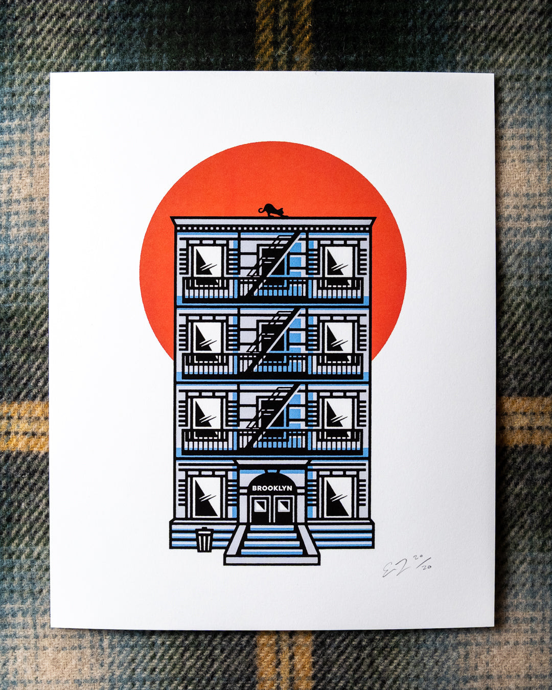 An 8"x10 art print featuring an illustrated NYC tenement building with a fire escape, black cat on the rooftop, BROOKLYN written above the doorway and a large orange-red sun behind it, photographed flat on a plaid fabric background.