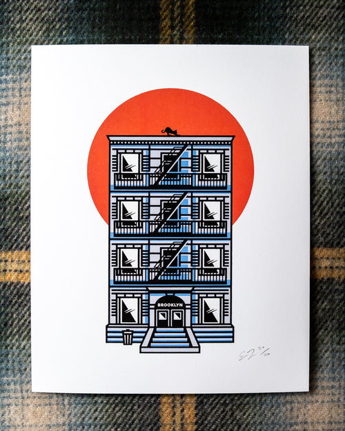 An 8"x10 art print featuring an illustrated NYC tenement building with a fire escape, black cat on the rooftop, BROOKLYN written above the doorway and a large orange-red sun behind it, photographed flat on a plaid fabric background.