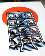 Close up of an 8"x10 art print featuring an illustrated NYC tenement building with a fire escape, black cat on the rooftop, BROOKLYN written above the doorway and a large orange-red sun behind it, photographed flat at an angle.