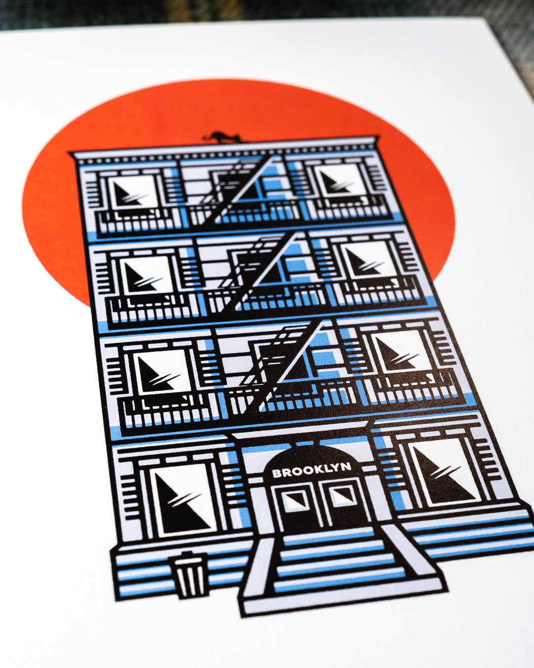 Close up of an 8"x10 art print featuring an illustrated NYC tenement building with a fire escape, black cat on the rooftop, BROOKLYN written above the doorway and a large orange-red sun behind it, photographed flat at an angle.