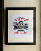 A framed 8"x10" white art print featuring a black-and-red illustrated storefront with the text 'Welcome To The Jerk Store' and 'You're Our All-Time Best Seller!', a reference to a quote by Jason Alexander's character George Costanza in the NBC sitcom Seinfield, placed on a plaid fabric background. Other details in the print include typography reading 'Shrimp Cocktail Served all day!' and Believe it or not, we're open!