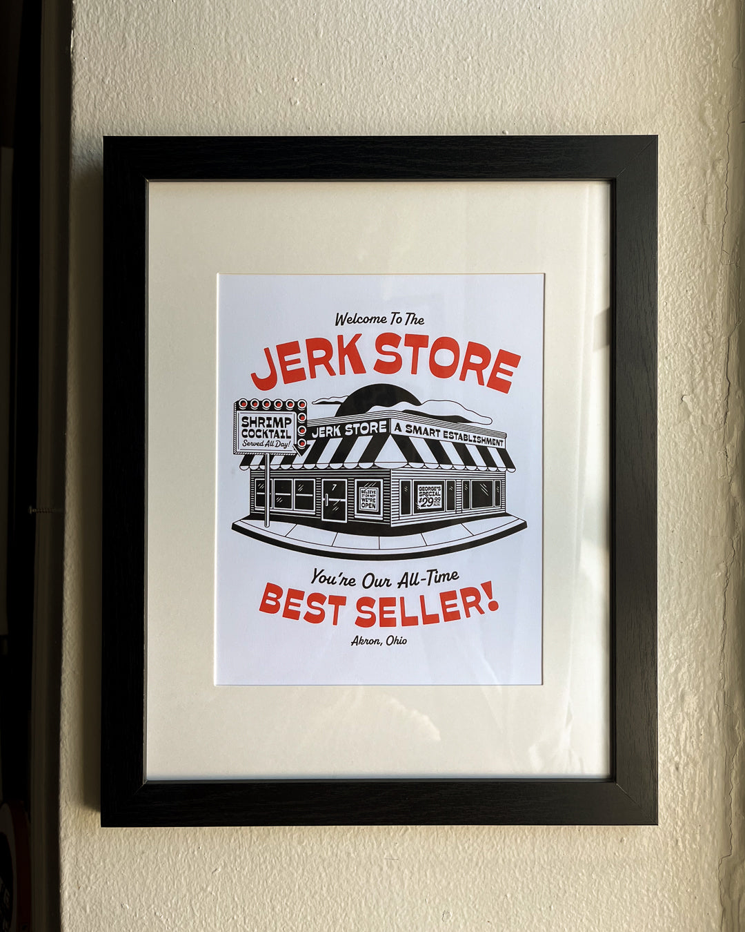 A framed 8"x10" white art print featuring a black-and-red illustrated storefront with the text 'Welcome To The Jerk Store' and 'You're Our All-Time Best Seller!', a reference to a quote by Jason Alexander's character George Costanza in the NBC sitcom Seinfield, placed on a plaid fabric background. Other details in the print include typography reading 'Shrimp Cocktail Served all day!' and Believe it or not, we're open!