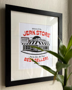A framed art print featuring a clean and bold illustration of a deli store with bold red text reading 'Welcome To The JERK STORE' and 'Best Seller!', displayed on a beige wall with a black frame and white mat. A green plant leaf partially obscures the lower right corner. Inspired by the character George Costanza from the NBC sitcom Seinfield.