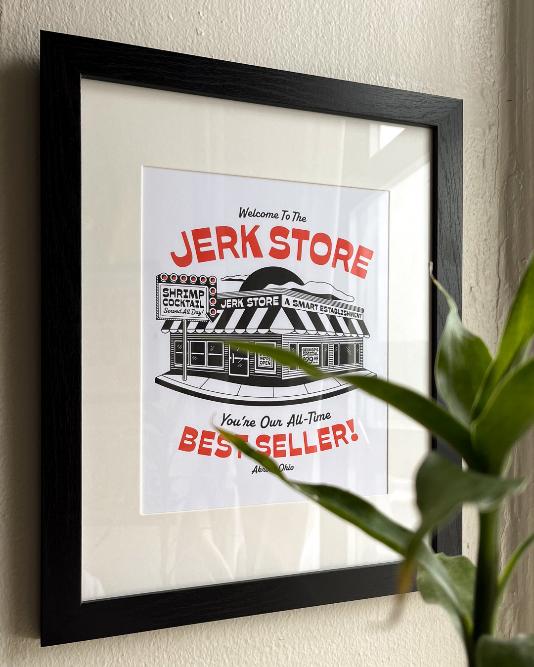 A framed art print featuring a clean and bold illustration of a deli store with bold red text reading 'Welcome To The JERK STORE' and 'Best Seller!', displayed on a beige wall with a black frame and white mat. A green plant leaf partially obscures the lower right corner. Inspired by the character George Costanza from the NBC sitcom Seinfield.