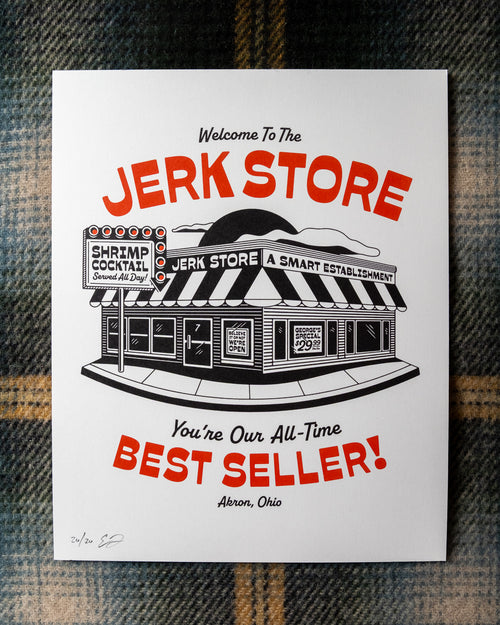 An 8"x10" white art print featuring a black-and-red illustrated storefront with the text 'Welcome To The Jerk Store' and 'You're Our All-Time Best Seller!', a reference to a quote by Jason Alexander's character George Costanza in the NBC sitcom Seinfield, placed on a plaid fabric background. Other details in the print include typography reading 'Shrimp Cocktail Served all day!' and Believe it or not, we're open!