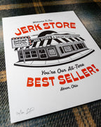 An angled shot of an 8"x10" white art print featuring a black-and-red illustrated storefront with the text 'Welcome To The Jerk Store' and 'You're Our All-Time Best Seller!', a reference to a quote by Jason Alexander's character George Costanza in the NBC sitcom Seinfield, placed on a plaid fabric background. Other details in the print include typography reading 'Shrimp Cocktail Served all day!' and Believe it or not, we're open!