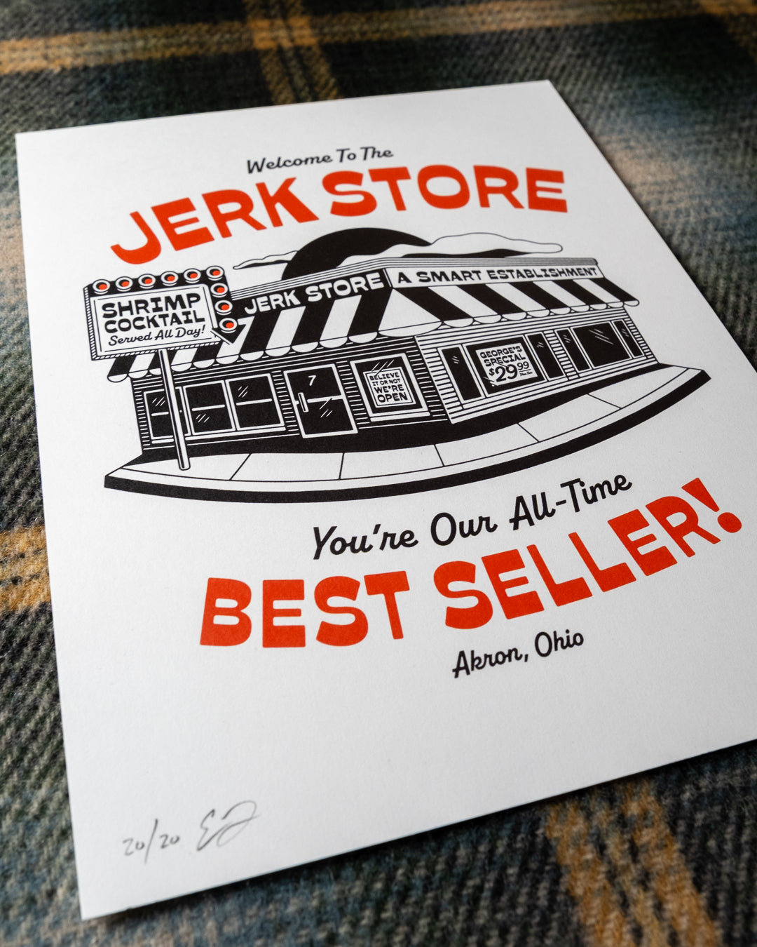 An angled shot of an 8"x10" white art print featuring a black-and-red illustrated storefront with the text 'Welcome To The Jerk Store' and 'You're Our All-Time Best Seller!', a reference to a quote by Jason Alexander's character George Costanza in the NBC sitcom Seinfield, placed on a plaid fabric background. Other details in the print include typography reading 'Shrimp Cocktail Served all day!' and Believe it or not, we're open!