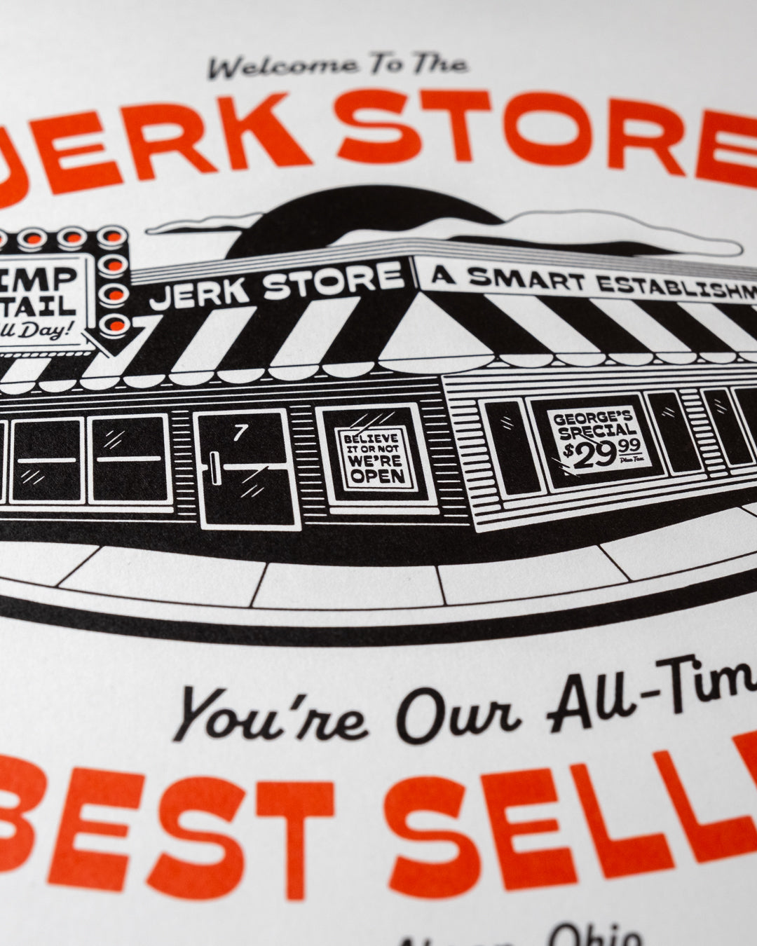 An close up shot of an 8"x10" white art print featuring a black-and-red illustrated storefront with the text 'Welcome To The Jerk Store' and 'You're Our All-Time Best Seller!', a reference to a quote by Jason Alexander's character George Costanza in the NBC sitcom Seinfield, placed on a plaid fabric background. Other details in the print include typography reading 'Shrimp Cocktail Served all day!' and Believe it or not, we're open!