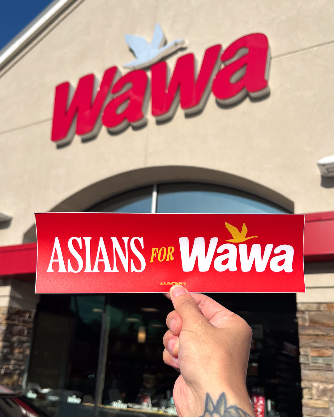 A hand holds a red bumper sticker that reads 'ASIANS FOR Wawa' in front of a Wawa store storefront in New Jersey or Philly with a large red logo on the beige building full of hoagies, soft pretzels and half gallons of iced tea. 