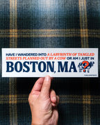 A hand holding a rectangular off-white bumper sticker with Boston Red Sox colors (blue and red) text that reads, 'HAVE I WANDERED INTO A LABYRINTH OF TANGLED STREETS PLANNED OUT BY A COW OR AM I JUST IN BOSTON, MA' with a cow-and-question-mark graphic. The background is a dark blue and gold plaid fabric.