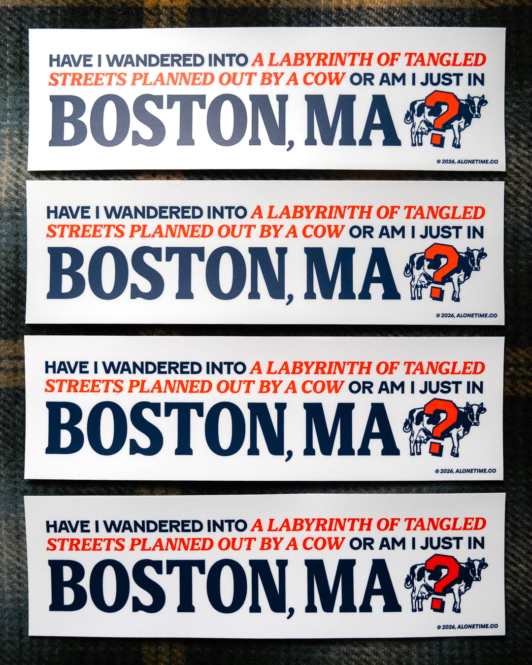 Four identical rectangular bumper stickers are stacked vertically on a dark fabric background. The stickers feature an off-white background with a humorous quote in red and navy blue (an ode to the Boston Red Sox) text, 'HAVE I WANDERED INTO A LABYRINTH OF TANGLED STREETS PLANNED OUT BY A COW OR AM I JUST IN BOSTON, MA', with 'BOSTON, MA' in large navy blue letters and an illustration of a cow with a question mark symbol over it.