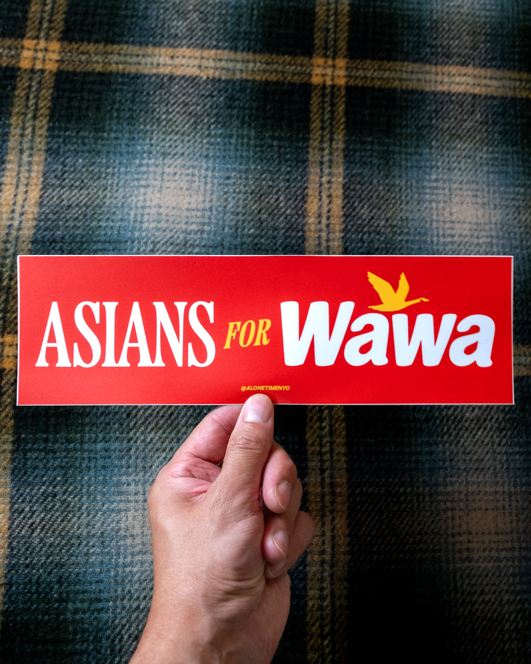 A hand holds a red rectangular bumper sticker that reads 'ASIANS FOR Wawa' with a yellow bird logo, set against a dark green and gold plaid fabric background.