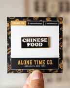 A hand holds a small enamel pin of an NYC Chinese food restaurant sign on a branded Alone Time Co. backing card. The pin reads “CHINESE FOOD” in white letters on a black background with a gold border.