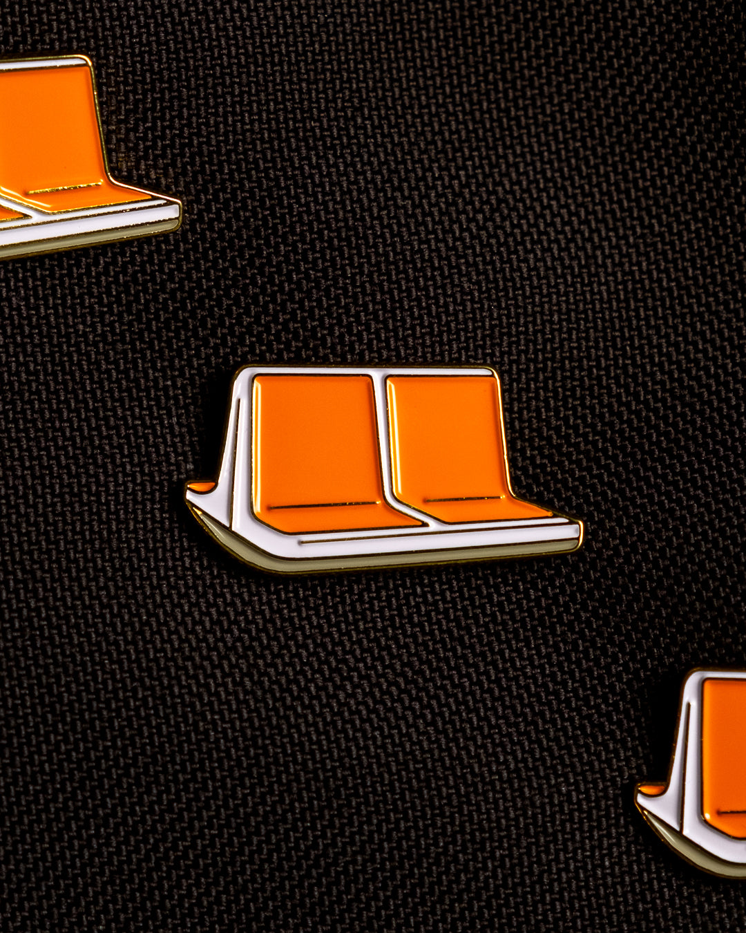 Three enamel pins shaped like classic NYC orange MTA subway seats with white sides and gold metal outlines pinned to a black canvas background