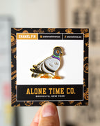 A close-up of a hand holding a card displaying an enamel pin shaped like a NYC pigeon wearing a beanie. The pin has white, black, gold, purple, and green detailing, mounted on a white and black card with 'ALONE TIME CO. Brooklyn, New York'.