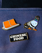 Three NYC inspired enamel pins displayed on a navy blue textured fabric background: an orange MTA subway seats pin, a pigeon wearing a beanie, and a black rectangular pin that reads 'CHINESE FOOD' in white letters.