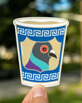 A die-cut sticker held by a person's fingers against a blurred outdoor background. The sticker depicts a stylized pigeon head inside a blue anthora cup-like border with a Greek key pattern.