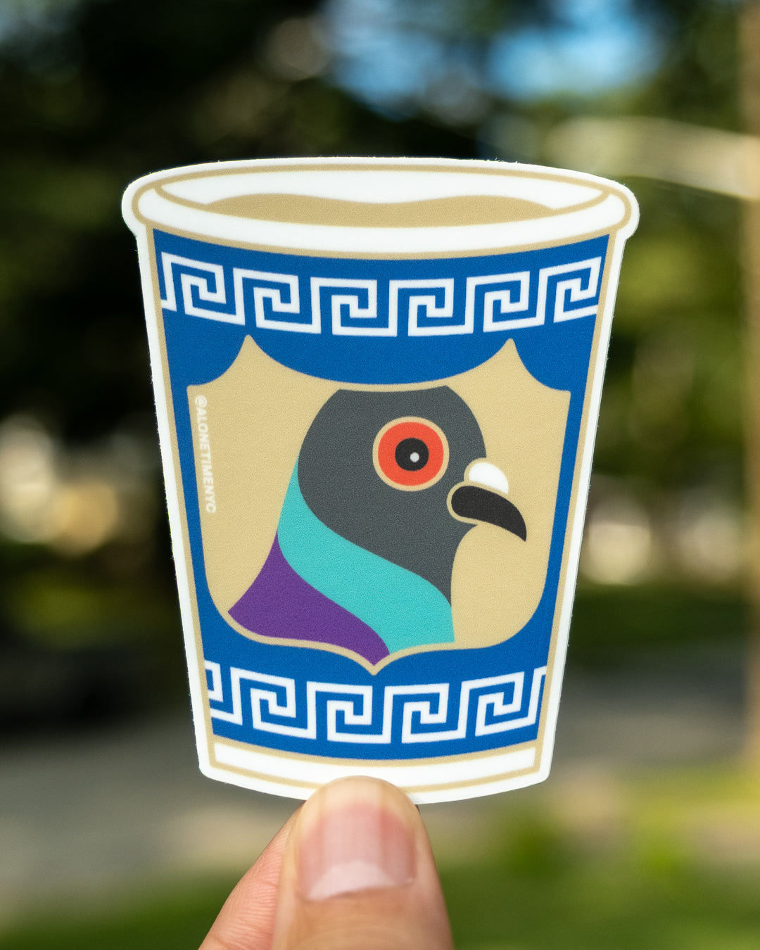 A die-cut sticker held by a person's fingers against a blurred outdoor background. The sticker depicts a stylized pigeon head inside a blue anthora cup-like border with a Greek key pattern.