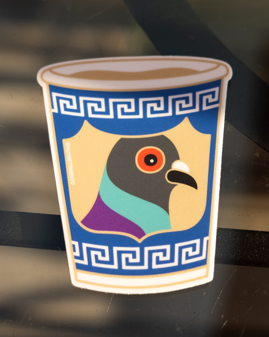 A die-cut sticker of an anthora cup featuring a stylized pigeon head illustration on a shield, bordered by blue Greek key patterns, shown against a dark reflective background.