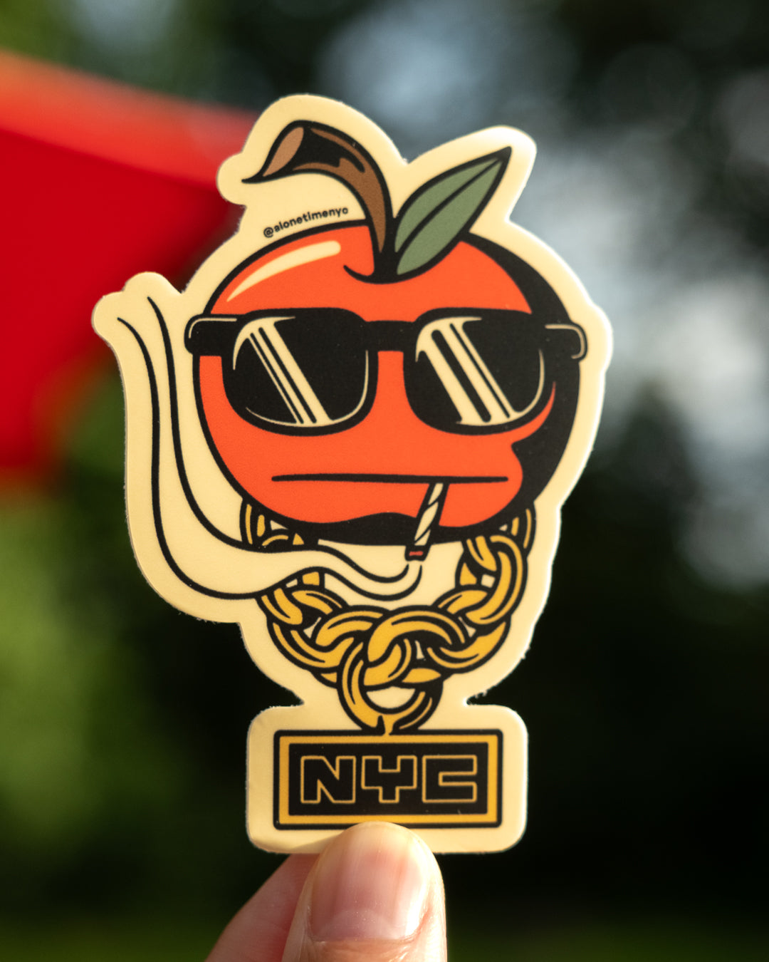 A die-cut sticker featuring a stylized red apple character wearing black sunglasses, a gold chain necklace, and a small 'NYC' plaque at the bottom, held by a hand against a blurred outdoor background.