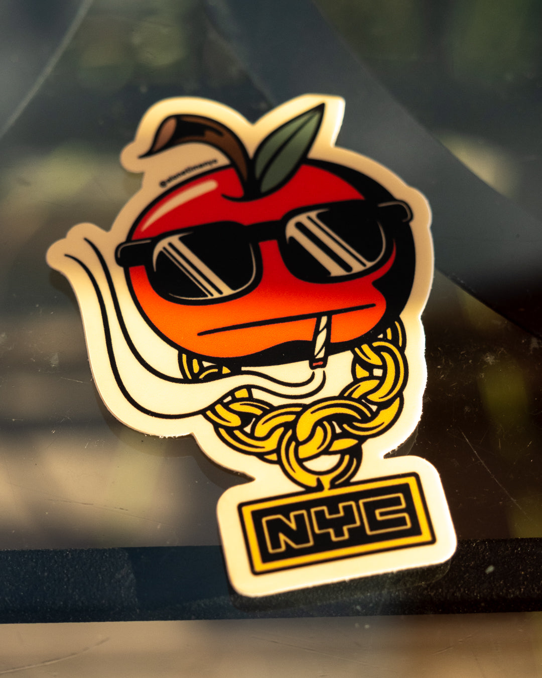 A die-cut sticker featuring a red illustrated cartoon apple wearing black sunglasses, a gold chain, and an NYC sign, shown close up on a dark reflective surface.