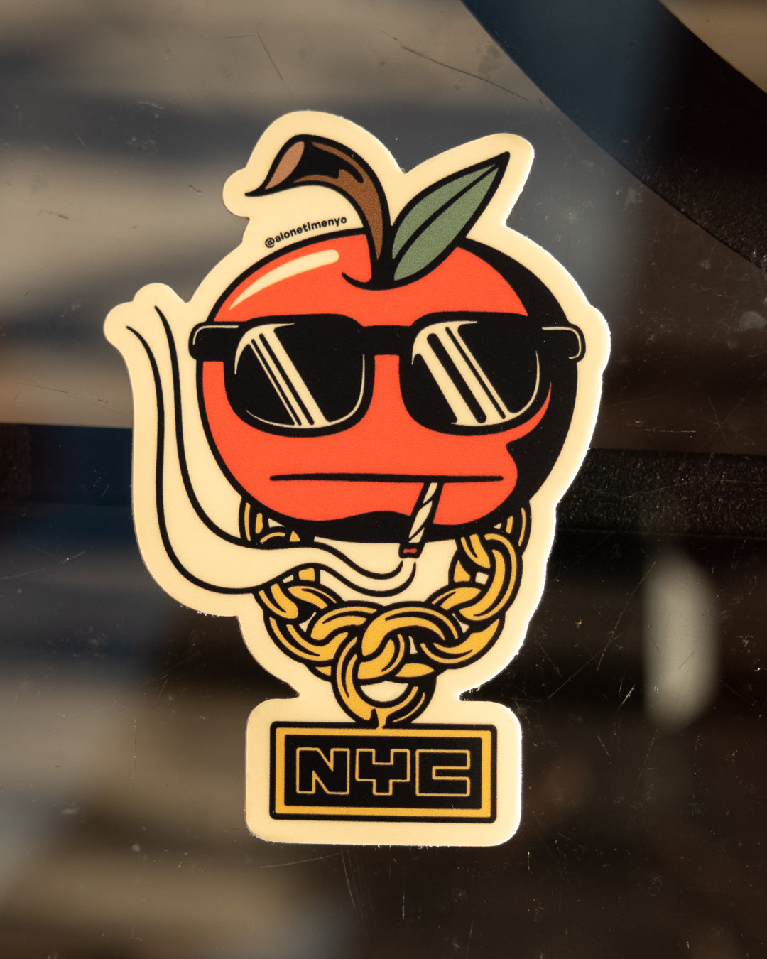 A die-cut sticker featuring a stylized red apple wearing black sunglasses, a gold chain necklace, and a small NYC sign below, shown on a dark reflective surface.