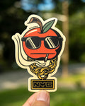 A die-cut sticker featuring a red "bad" apple character wearing black sunglasses, a gold NYC chain, smoking a joint, held by a hand against a blurred outdoor background. The sticker has a cream border and an 'NYC' box at the bottom.