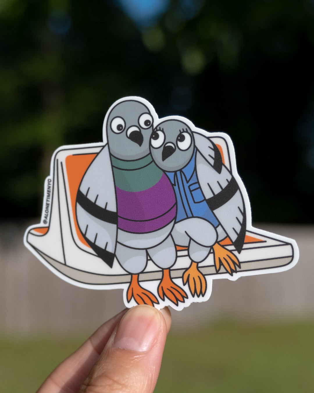 A die-cut vinyl sticker of two illustrated cartoon pigeons lovingly staring at each other and sitting on an NYC MTA classic orange subway seat, held by a hand against a blurred outdoor background.