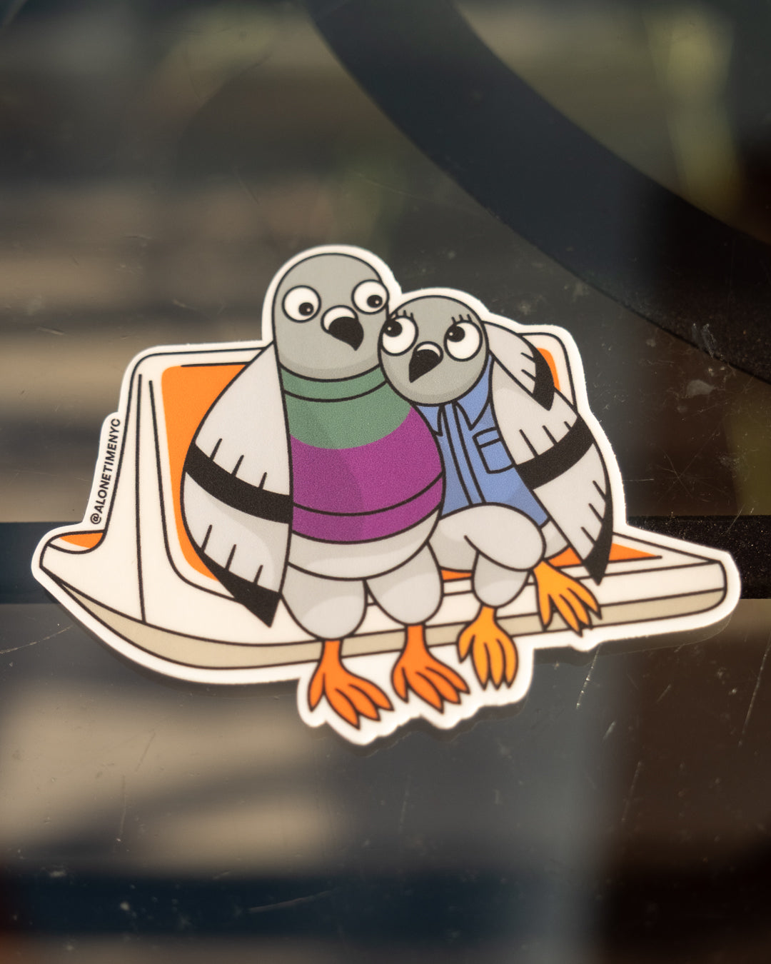 A die-cut vinyl sticker featuring two illustrated / cartoon pigeons, wearing a sweater and a button up shirt, sitting on an NYC MTA classic orange subway seat, shown applied to a dark glass surface. The sticker has a white border and includes the instagram handle @alonetimenyc printed vertically on the left side of the seat.