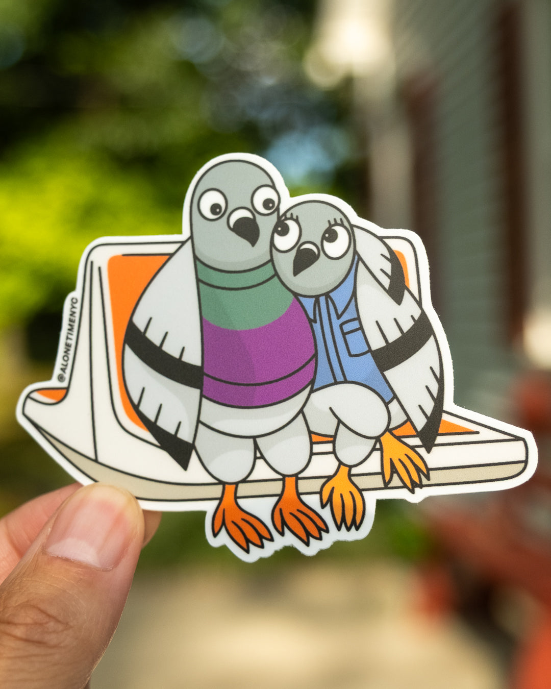 A die-cut sticker featuring two illustrated / cartoon pigeons, wearing a sweater and button up shirt, sitting on a classic NYC MTA orange subway seat, held by a hand against a blurred outdoor background. The sticker has a white border and shows the instagram handle '@alonetimenyc' printed vertically on the left side.