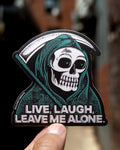 A die-cut sticker featuring a heavily stippled illustration of a grim reaper skull in a green hood, holding a sickle, with the text 'LIVE, LAUGH, LEAVE ME ALONE.' A human hand is holding the sticker against a blurred outdoor background.