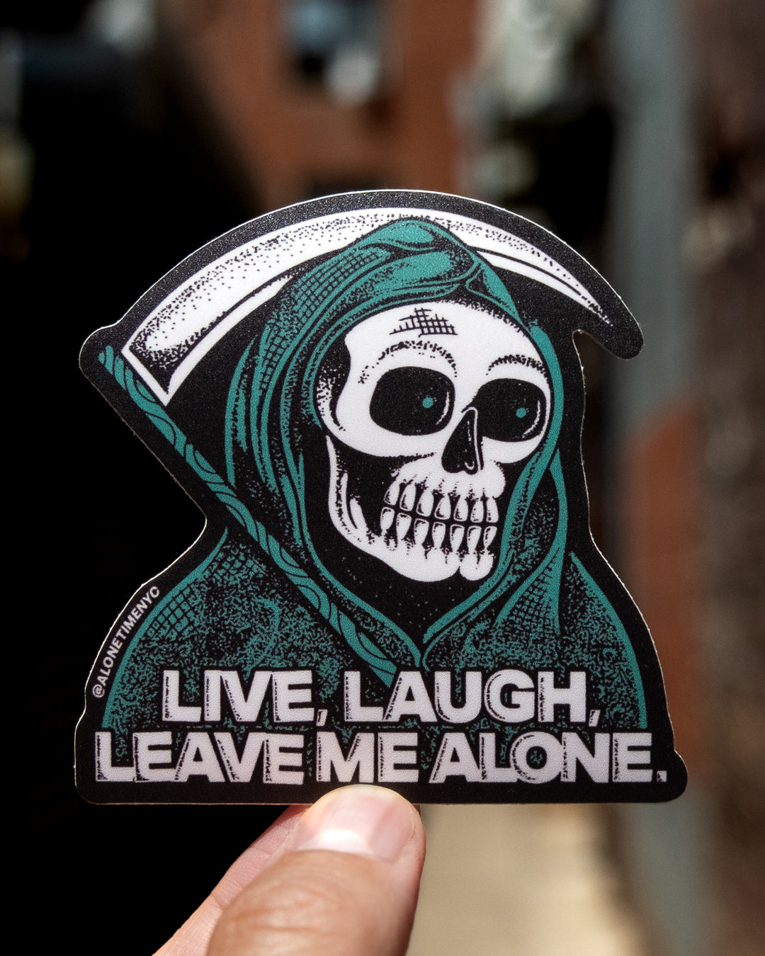 A die-cut sticker featuring a heavily stippled illustration of a grim reaper skull in a green hood, holding a sickle, with the text 'LIVE, LAUGH, LEAVE ME ALONE.' A human hand is holding the sticker against a blurred outdoor background.