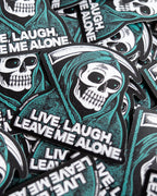 A pile of die-cut stickers featuring a heavily stippled illustration of a grim reaper skull in a green hood, holding a sickle, with the text 'LIVE, LAUGH, LEAVE ME ALONE.' 