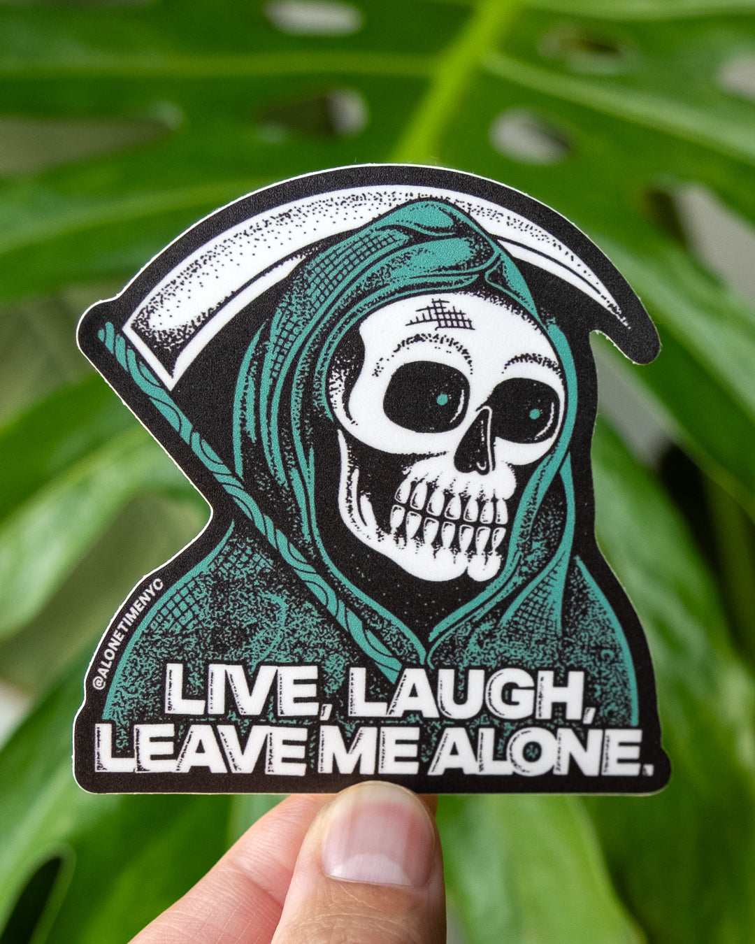 A die-cut sticker featuring a grim reaper skull, holding a sickle, illustration with the text 'LIVE, LAUGH, LEAVE ME ALONE.' A hand is holding the sticker in front of large green monstera leaves.