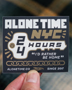 Die-cut vinyl Sticker with 'Alone Time NYC 24 Hours' and 'I'd rather be home' text and an illustration of a house key and gold chain links, held by a hand against a blurred background.