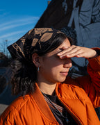 Black screen printed bandana with gold rose illustrations, chain links and 'Alone Time' typography, worn by a person wearing an orange jacket in front of a blurred Brooklyn background.