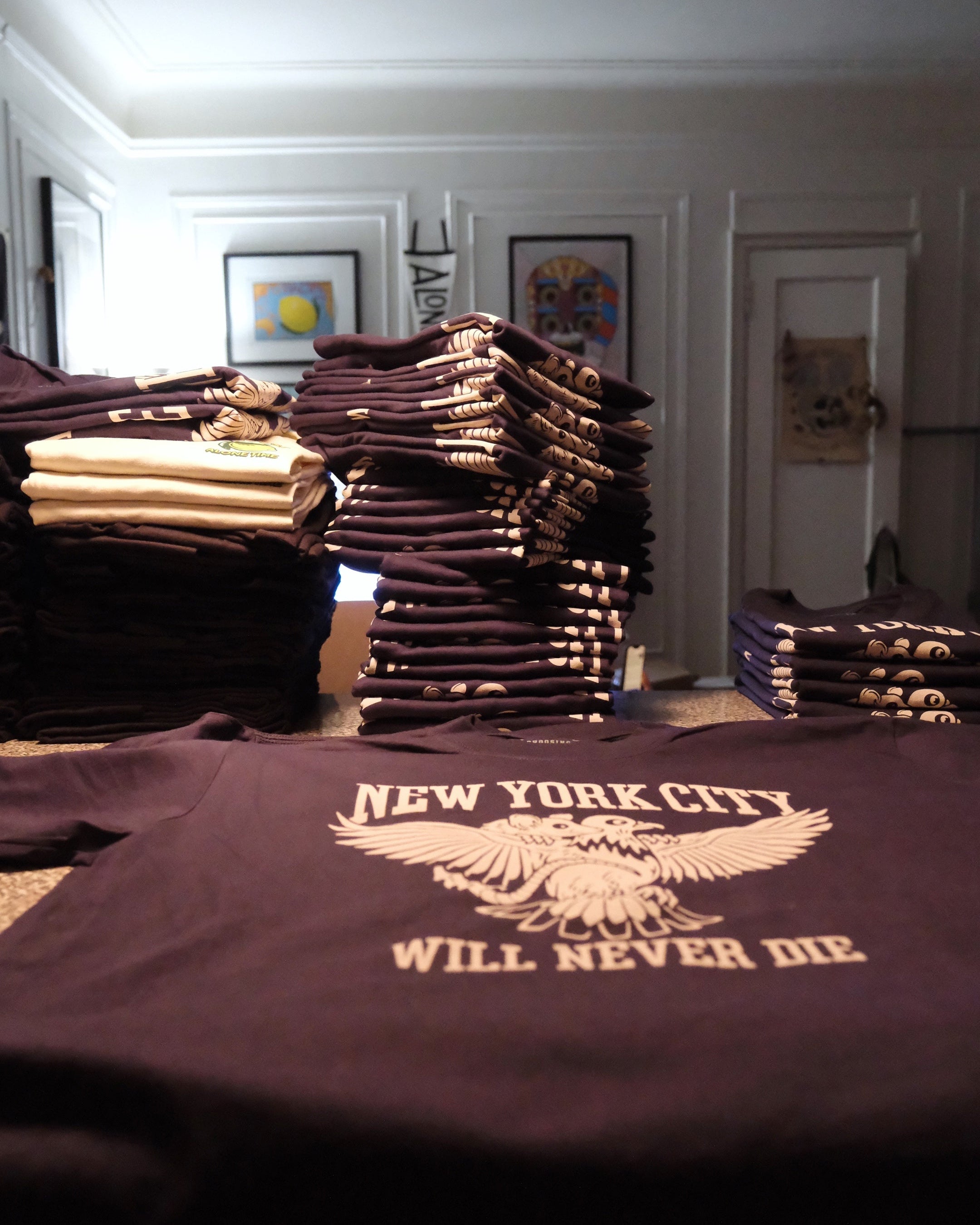 Stacks of t-shirts with 'New York City Will Never Die' design on a table.